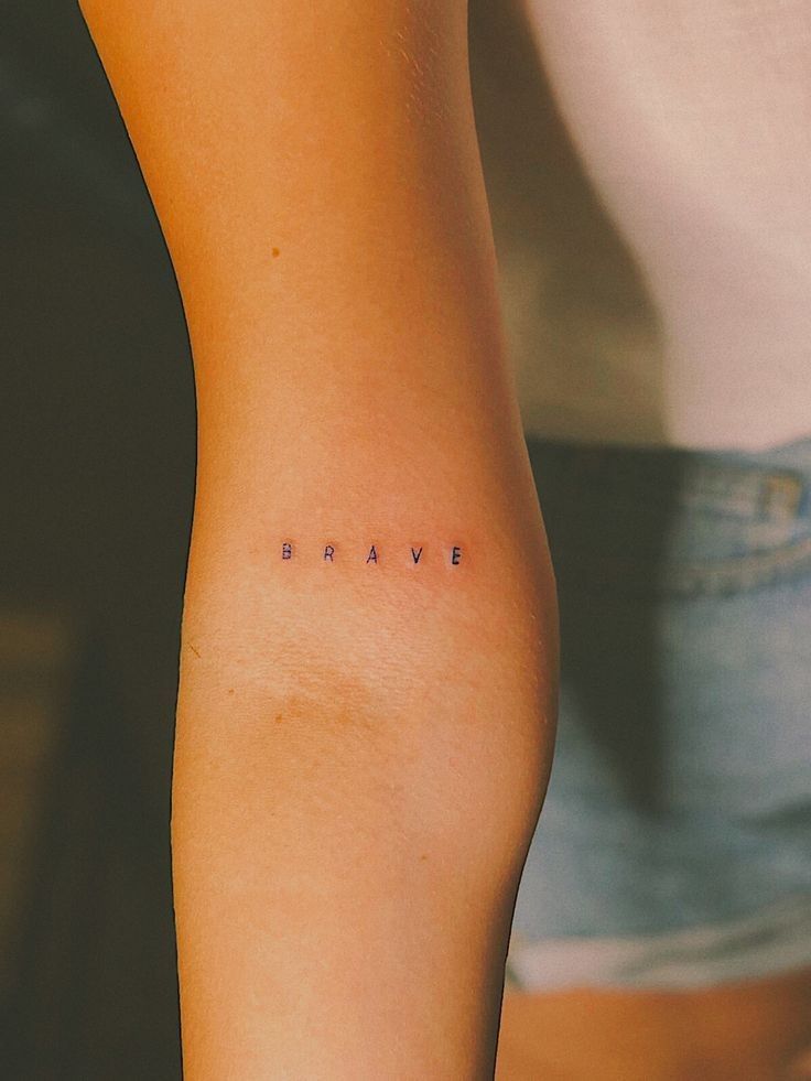 tatouage minimaliste en lettrines "BRAVE" sur l'avant-bras d'une femme